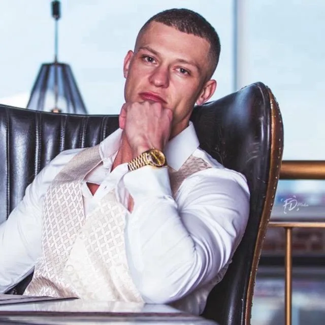 Young man in a white shirt and beige vest sitting on a black leather chair indoors.