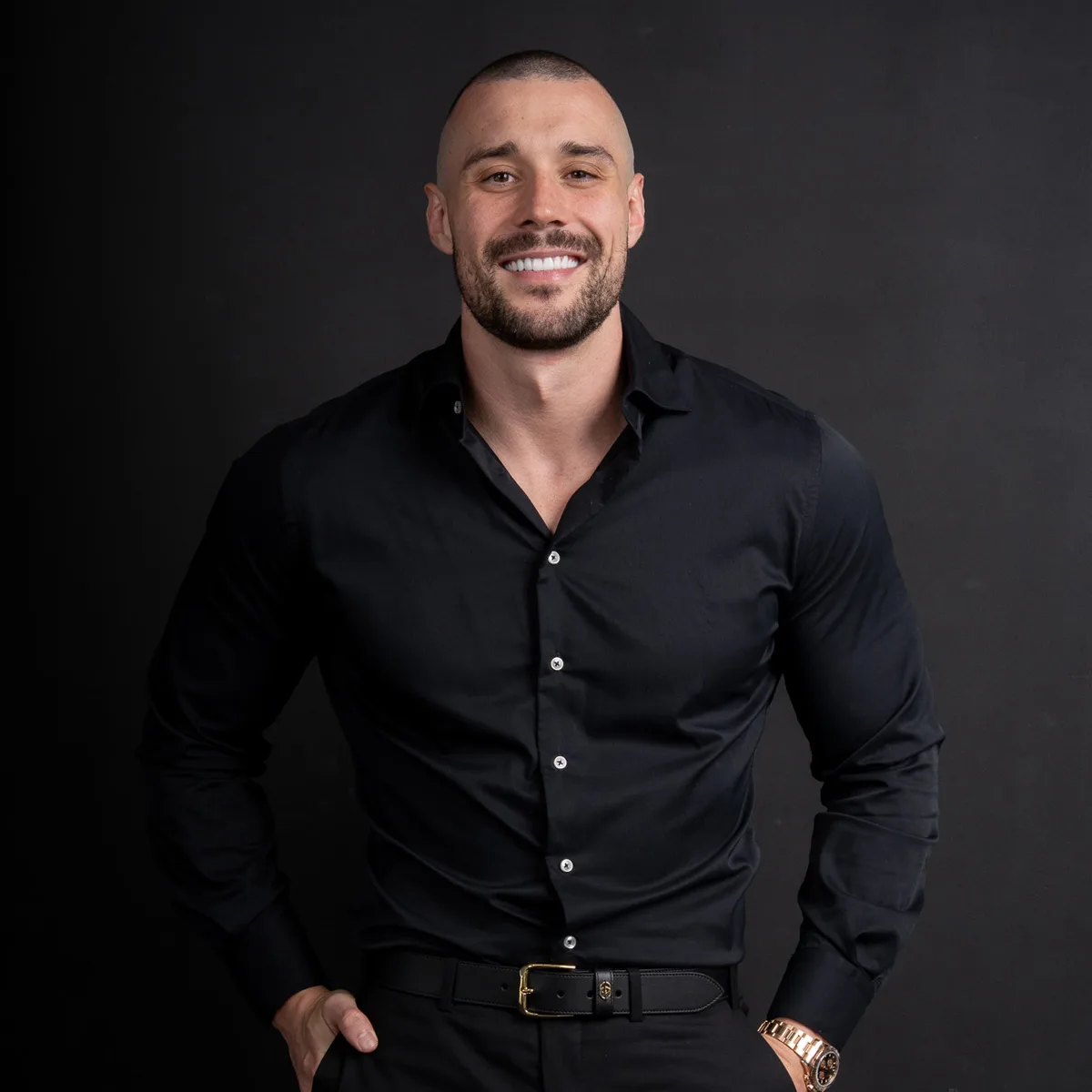 Smiling man with short hair and beard wearing a black button-up shirt and gold watch on black background.