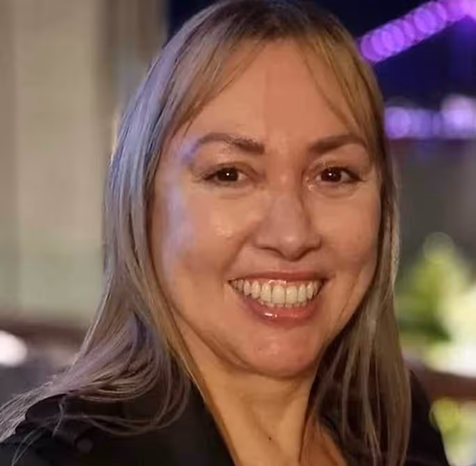 Smiling woman with long blonde hair wearing a black top in a softly lit indoor setting.