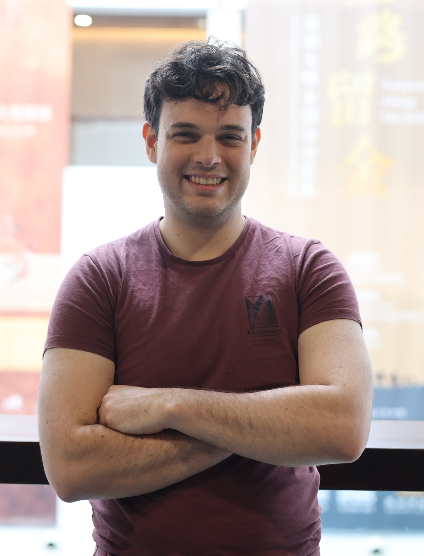 Smiling young man with curly dark hair in a maroon T-shirt crosses his arms indoors.