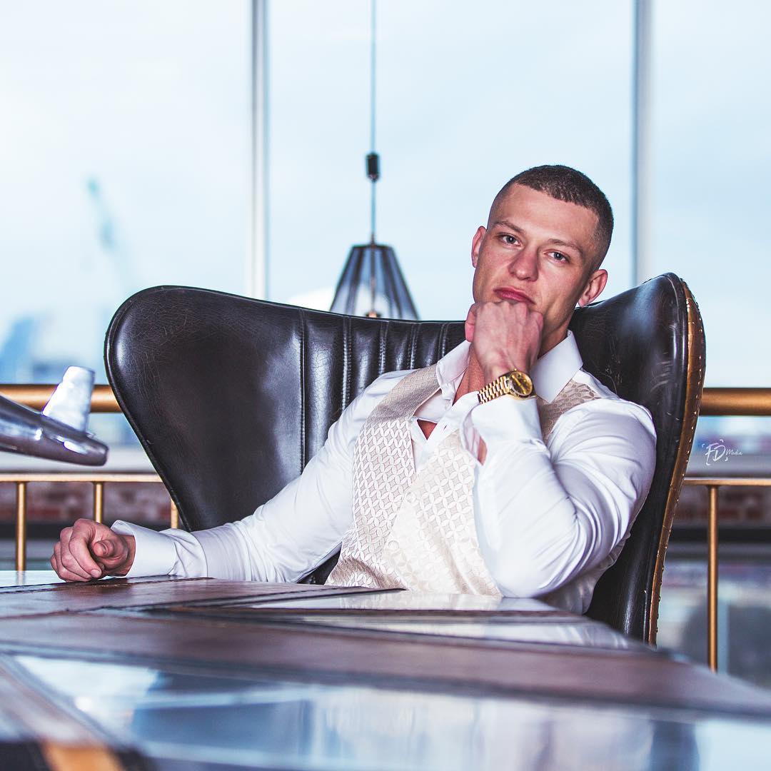 Man in white shirt and patterned vest sitting in a black leather chair at a table, resting chin on fist.